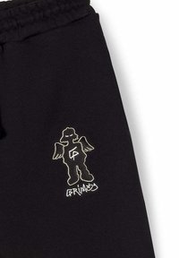 Pantalones de chándal negros con un personaje de dibujos animados bordado en blanco, con alas de ángel y texto, creando un detalle visual contrastante.