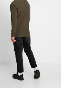 Camicia a maniche lunghe color oliva con orlo arrotondato, abbinata a jeans scuri. Il modello indossa sneakers nere con calze bianche, visto da dietro.