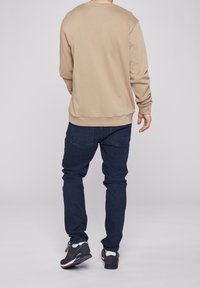 Beige sweatshirt med långa ärmar och rund hals, kombinerad med mörkblå jeans och svarta träningsskor med röda detaljer.