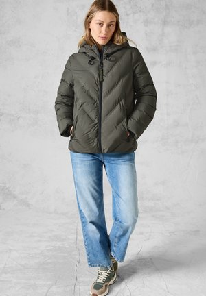 Doudoune vert foncé avec capuche, présentant un motif en chevrons, fermeture éclair et accents avec cordon de serrage. Portée avec un jean en denim clair et des baskets.