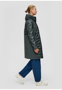 Lang donkergrijze jas met een puffy, gewatteerde textuur aan de bovenkant. Heeft een capuchon, zijzakken en relaxte blauwe jeans met witte sneakers.