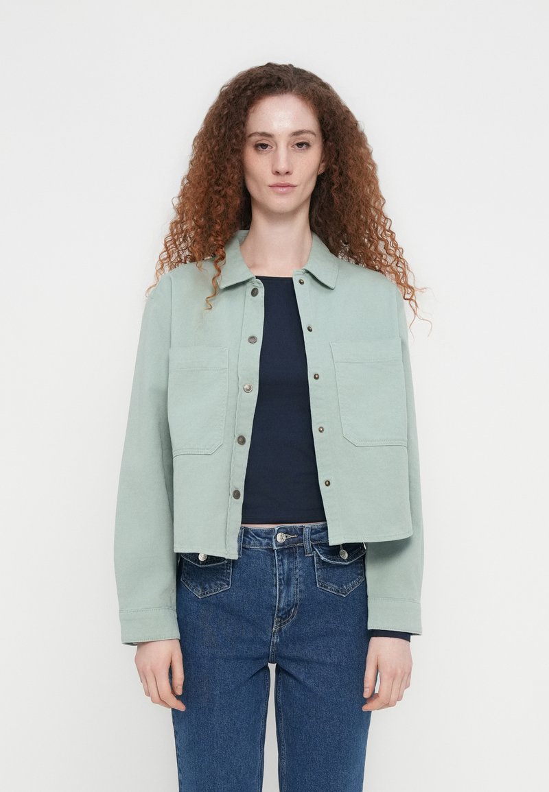 Veste courte vert menthe en tissu texturé. Comprend un col pointu, deux poches poitrine et des boutons-pression argentés. Portée par-dessus un haut foncé.