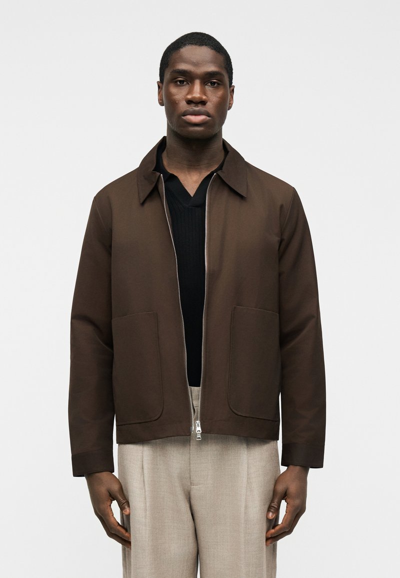 Άνδρας που φοράει καφέ ζακέτα με φερμουάρ πάνω από μαύρο πουκάμισο και ανοιχτό μπεζ παντελόνι, στέκεται μπροστά σε ένα απλό λευκό φόντο.