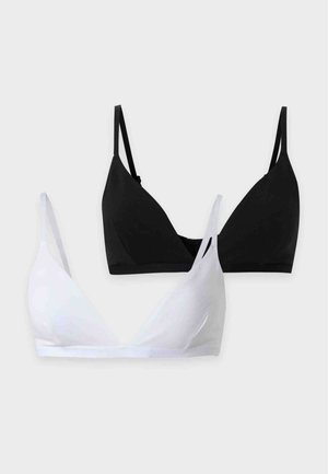 Femme debout pieds nus portant un bikini noir à fines bretelles sur fond blanc uni.