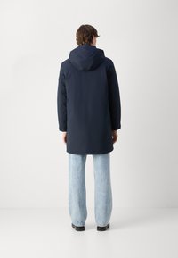 Mørkeblå parka med hætte, fremstillet af et glat, vandafvisende stof. Båret over lyseblå, løstsiddende jeans og sorte sko.