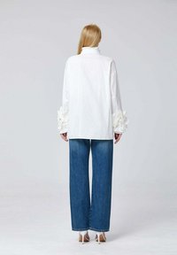 Haut blanc à manches longues avec des manches volantes, associé à un pantalon en denim bleu à jambes larges. Le mannequin porte des chaussures à talons beiges.