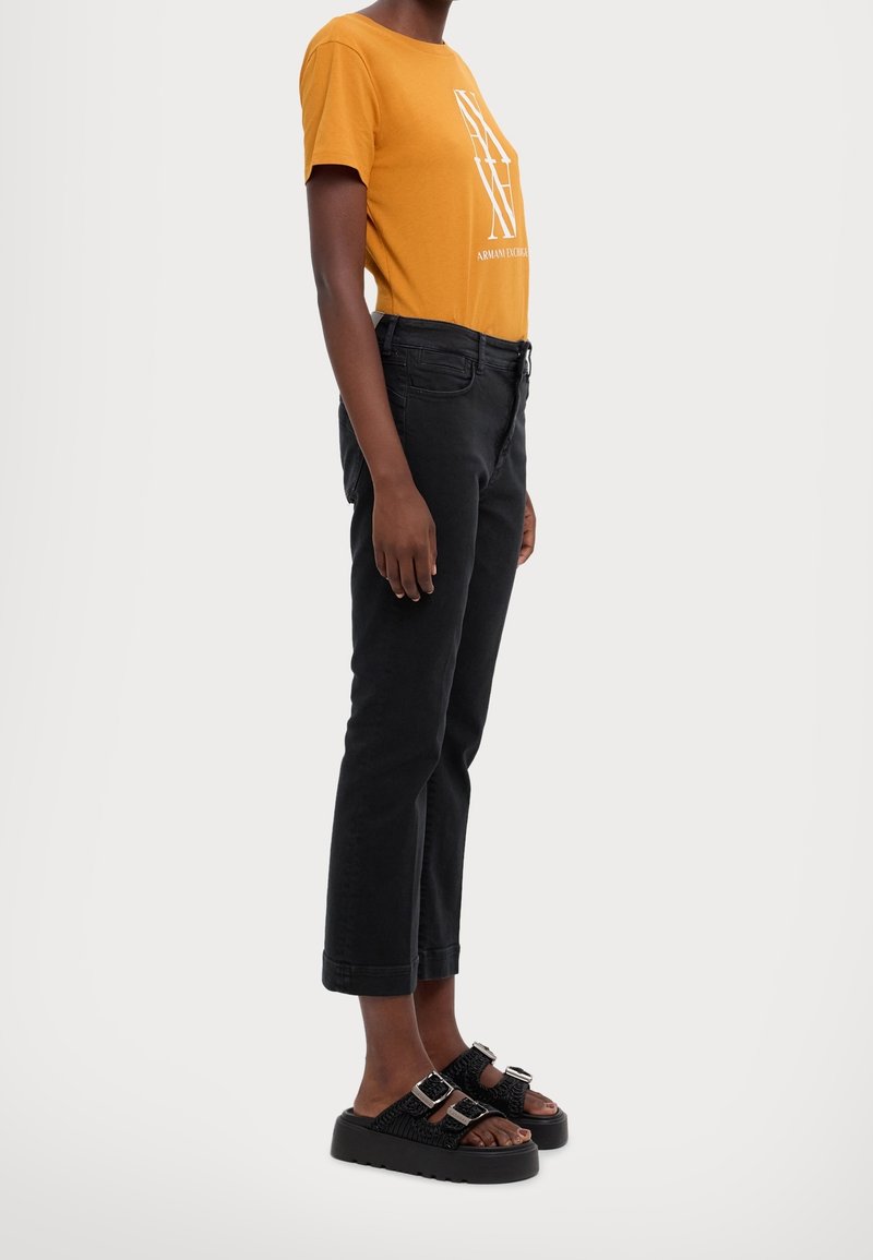 Mustard yellow t-shirt with white logo, black slim-fit jeans, and chunky black sandals with double buckle accents.