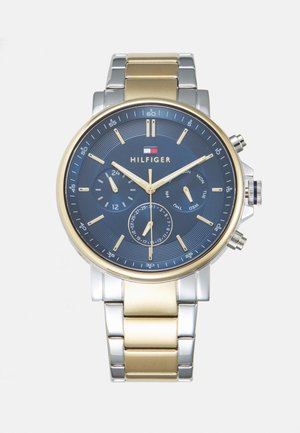 Sudraba un zelta toņu metāla pulkstenis ar zilu teksturētu ciparnīcu, hronogrāfa funkcijām, zelta rādītājiem un vairākiem subciparnīcām. Hilfiger logo.