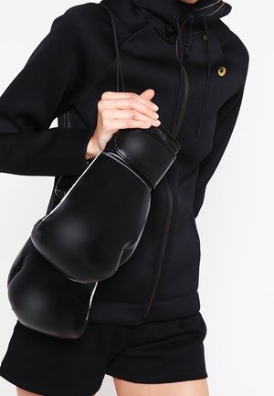 Personne en sweat à capuche noir et short tenant de grandes gants de boxe noirs par leurs sangles sur un fond blanc.