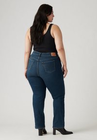 Tamsiai mėlynos džinsinės kelnės su vidutiniu liemens aukščiu, dviem užpakalinėmis kišenėmis ir odiniu prekės ženklo ženklu. Dėvimos su juodu berankoviu marškinėliu ir juodais aukštakulniais batais.