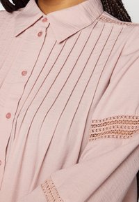 Rosa Hemd mit Knöpfen, das aus einem strukturierten Stoff gefertigt ist, vertikale Falten und Spitzenakzente an den Ärmeln für zusätzlichen Detailreichtum und Design aufweist.