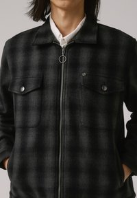 Veste à carreaux noirs en tissu à texture brossée. Elle possède une fermeture éclair à l'avant, deux poches poitrine et des boutons-pression en argent.