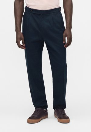 Man draagt donkerblauwe broek en donkerbruine sneakers met rubberen zolen, staand tegen een effen lichte achtergrond.