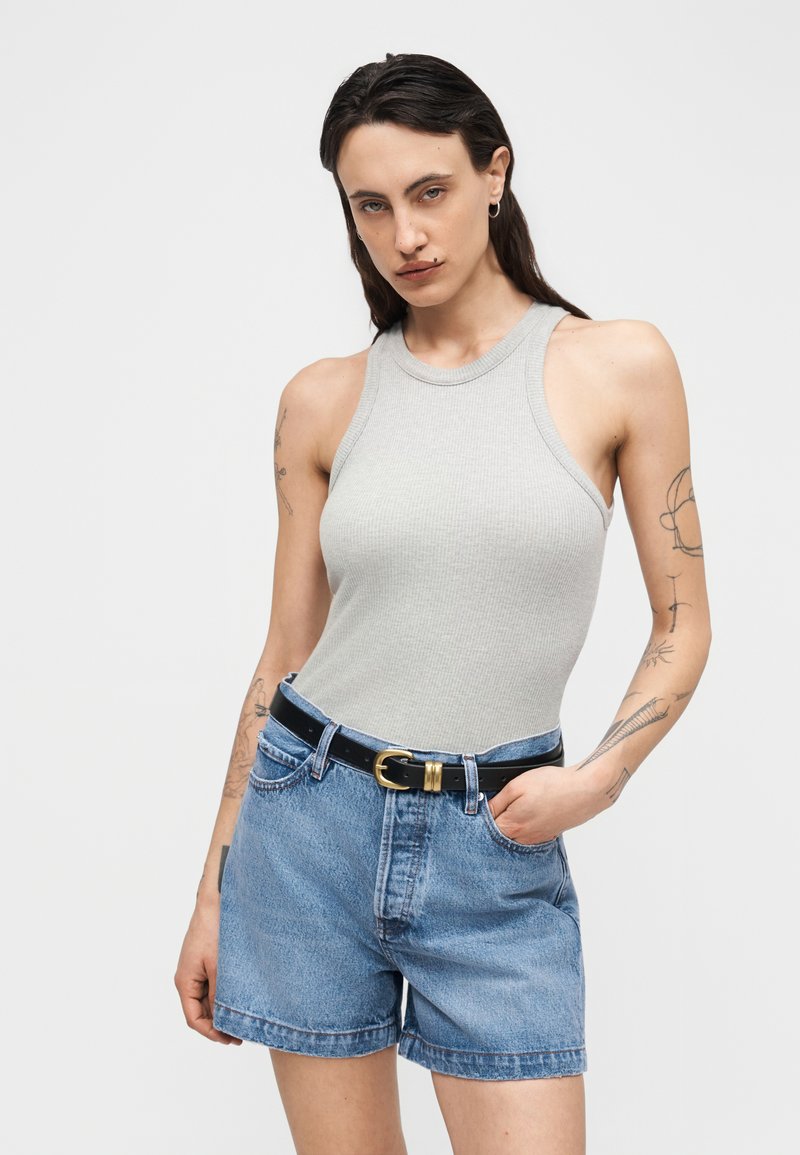 Frau trägt ein hellgraues, ärmelloses, geripptes Oberteil, hoch taillierte Jeansshorts mit einem schwarzen Gürtel und Tattoos auf beiden Armen.