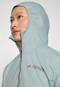 Lightweight, mint green jacket with grid pattern, high collar, and adjustable hood. Features a brown zipper and TERREX branding.