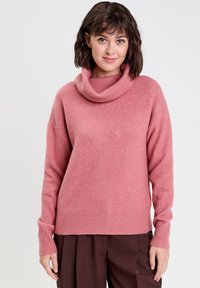 Pull en col roulé rose en matériau doux et texturé. Présente des poignets et un ourlet côtelés, avec une coupe décontractée et des épaules basses.