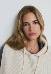 Hellbeige, gestrickter Pullover mit Kapuze. Verfügt über einen strukturierten Stoff und einen Kordelzug. Über einem grauen Oberteil getragen, mit glattem, welligem Haar.