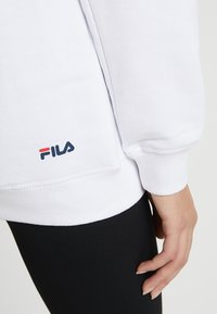 Felpa bianca con polsini e orlo a costine, caratterizzata da un piccolo logo FILA blu e rosso in basso a sinistra. Indossata sopra leggings neri.