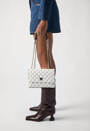 Quilted white handbag with black accents, silver chain straps, and a triangular logo, elegantly held alongside knee-high brown boots.