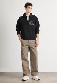 Musthave musta tõmblukuga kapuutsiga sviiter CK logo kujutisega, koos lõtvade beežide pükstega ja valgete tosadega. Sile kangas ja struktureeritud disain ribikääridega.
