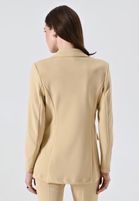 Blazer beige su misura con una texture liscia, maniche lunghe e una silhouette aderente. Presenta un collo classico e dettagli di cucitura sul retro.