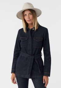 Giacca in denim scuro con vita cintata, due tasche sul petto e cuciture arancioni a contrasto. Abbinata a un cappello a tesa larga chiara.