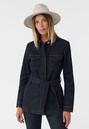 Dark denim jacket with a belted waist, two chest pockets, and contrasting orange stitching. Paired with a light wide-brimmed hat.
