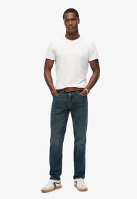 Camisa de algodón blanca, jeans de mezclilla azul, cinturón marrón, zapatillas blancas con acentos en gris y negro. Modelo de pie con las manos en los bolsillos.