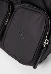 Zaino in nylon nero con due tasche con zip, logo stampato e dettagli in metallo argentato. Texture liscia con cuciture resistenti.