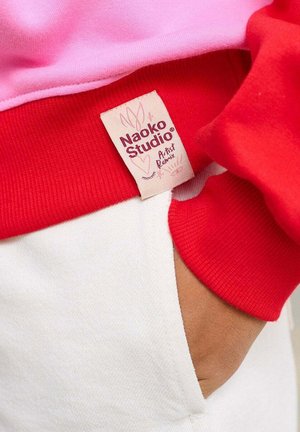 Czerwona i różowa bluza z prążkowanymi mankietami i białą kieszenią. Etykieta z napisem „Naoko Studio” i kwiatowym wzorem. Widoczna miękka struktura.
