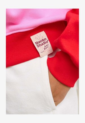 Czerwona i różowa bluza z prążkowanymi mankietami i białą kieszenią. Etykieta z napisem „Naoko Studio” i kwiatowym wzorem. Widoczna miękka struktura.