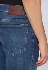 Jeans de mezclilla azul desgastados con un parche de cuero marrón que muestra "TRUSSARDI". Presentan un diseño clásico de cinco bolsillos y detalles de costura visibles.
