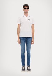 White polo shirt with black trim, worn with blue skinny jeans and dark sneakers. Eyewear is black and rectangular. Simple, casual attire.