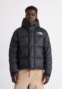 Svart pufferjacka med hög krage, quiltat mönster och The North Face-logotyp på bröstet. Har dragkedjefickor och elastiska ärmmuddar.