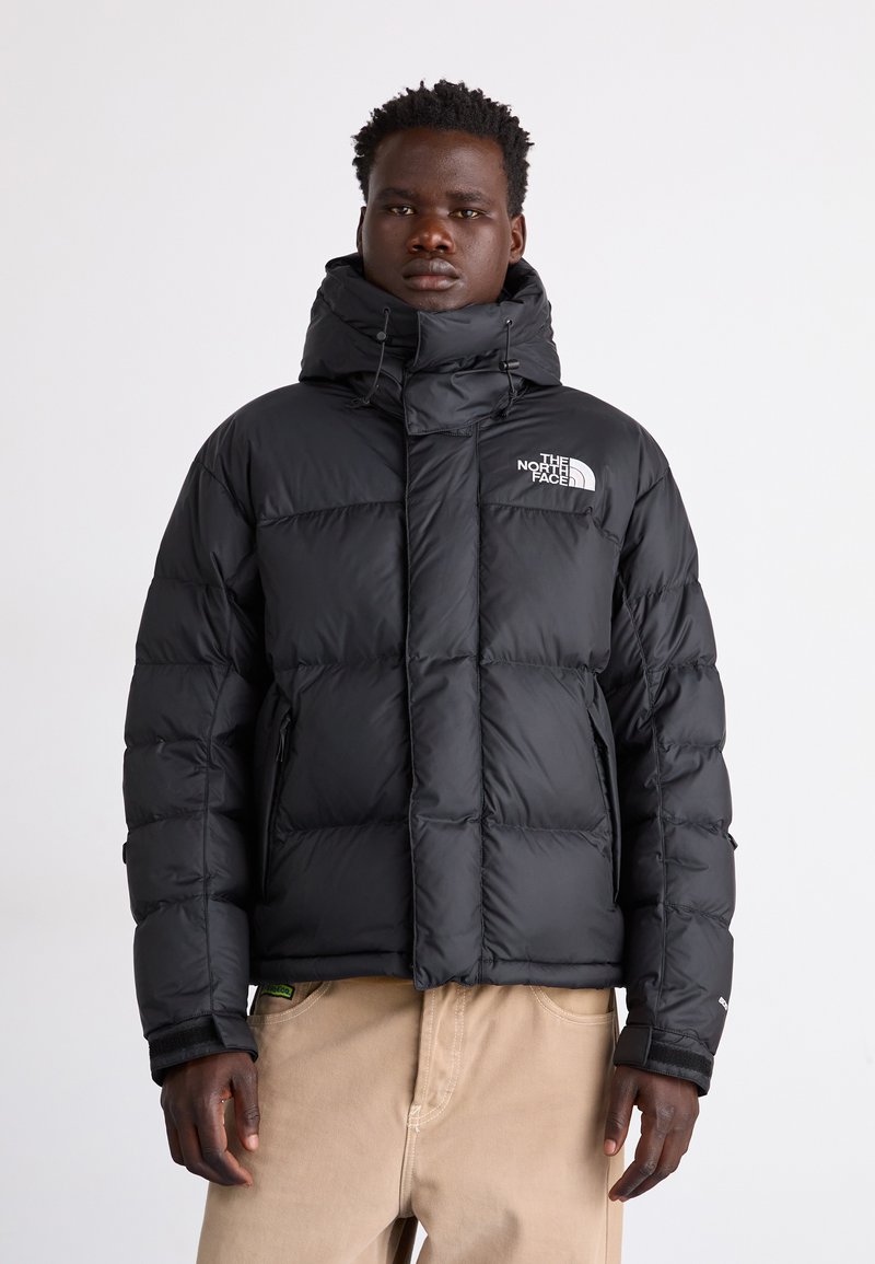 Svart pufferjacka med hög krage, quiltat mönster och The North Face-logotyp på bröstet. Har dragkedjefickor och elastiska ärmmuddar.
