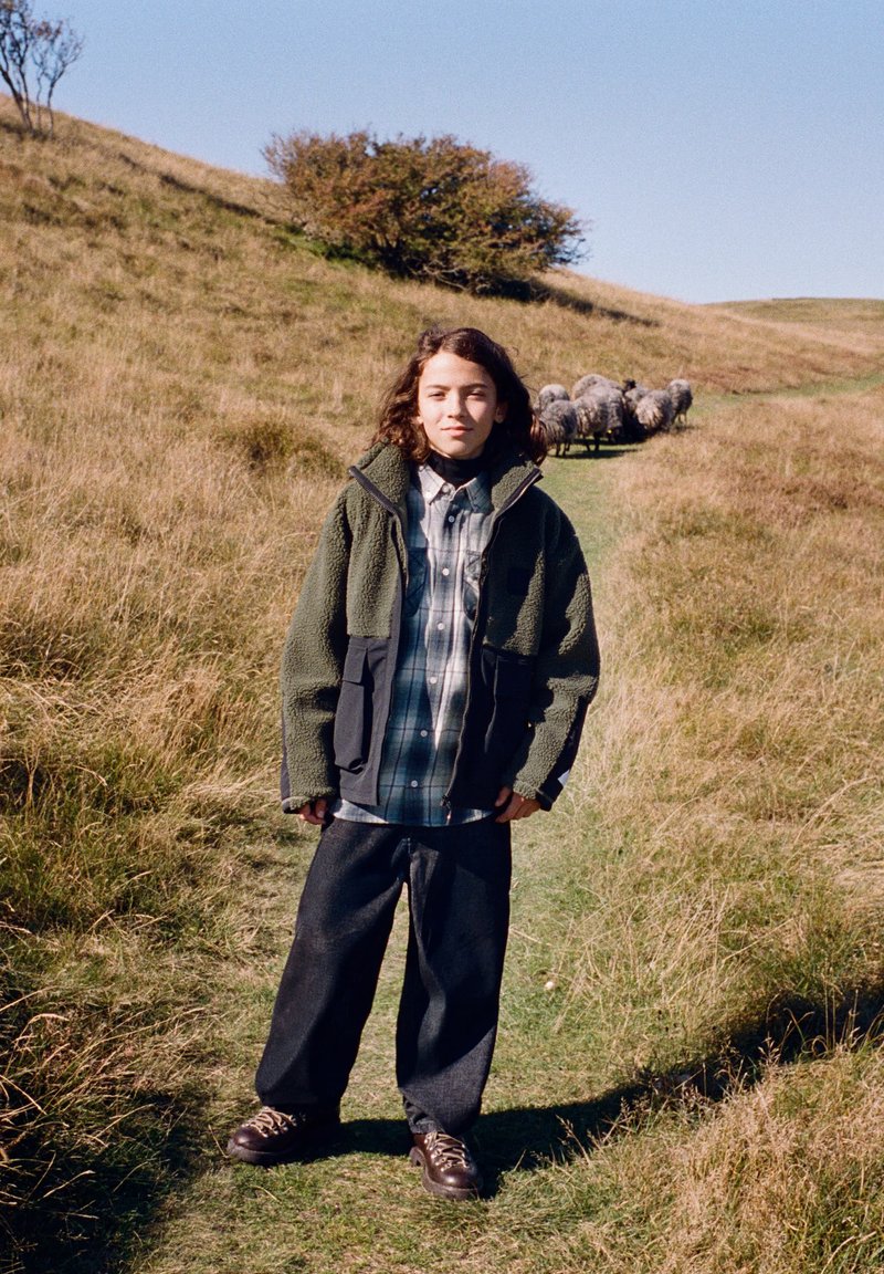 Jeune personne en tenue d'extérieur debout sur un sentier herbeux avec un troupeau de moutons marchant derrière sur une colline sous un ciel clair.