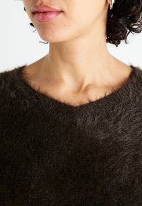 Brauner, flauschiger Pullover mit V-Ausschnitt. Weiches, strukturiertes Material und leicht lockere Passform. Nahaufnahme des Ausschnitts und der Haut.