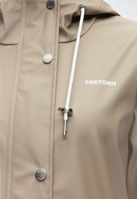 Veste de pluie beige avec cordon de serrage blanc, boutons-pression en métal gravés "Tretorn Sweden" et logo "TRETORN" sur la poitrine.