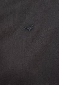 Petit logo de cheval brodé en noir, centré sur un fond de tissu gris foncé.