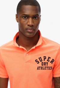 Koraloowa koszulka polo z kołnierzykiem, z haftowanym na czarno logo "SUPER DRY ATHLETICS" po lewej stronie. Miękka faktura z krótkimi rękawami.