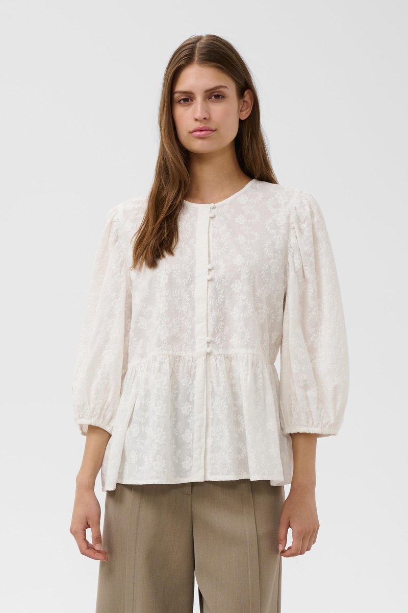 Jeune femme portant un chemisier en dentelle blanche à motif floral avec des manches trois-quarts bouffantes et un pantalon ample beige sur un fond uni.