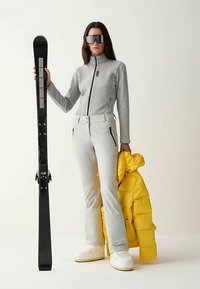 Ski outfit featuring a gray zip-up top, light gray pants with zippers, white boots, and a bright yellow puffer jacket, holding a black ski.