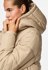 Lys beige puffjakke med høy krage og hette. Har ribbede mansjetter og sydd detaljer på stoffet. Glatt tekstur.