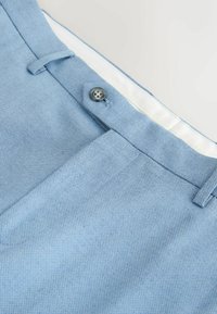 Ceinture de pantalon en tissu bleu clair texturé avec passants pour ceinture, fermeture par bouton, et partie de la zone de la fermeture éclair visible sur un fond blanc.