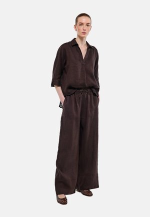 Femme debout portant une chemise marron foncé à col en V ample avec les manches retroussées, un pantalon à jambes larges et des chaussures tissées plates, les mains dans les poches.