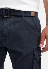 Man wearing navy blue cargo shorts with a black belt and a white shirt, hand relaxed by side.