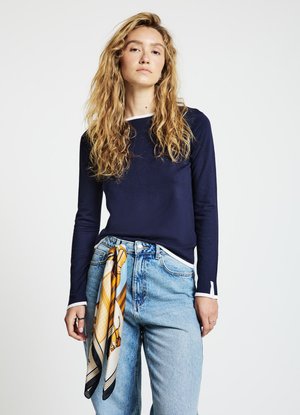 Haut à manches longues bleu marine avec des accents blancs, associé à un jean taille haute bleu clair et une écharpe à motifs nouée à la taille.