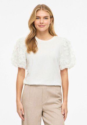 Donna bionda che indossa una blusa bianca con maniche a sbuffo e motivo floreale e pantaloni beige a vita alta, in piedi su sfondo bianco.