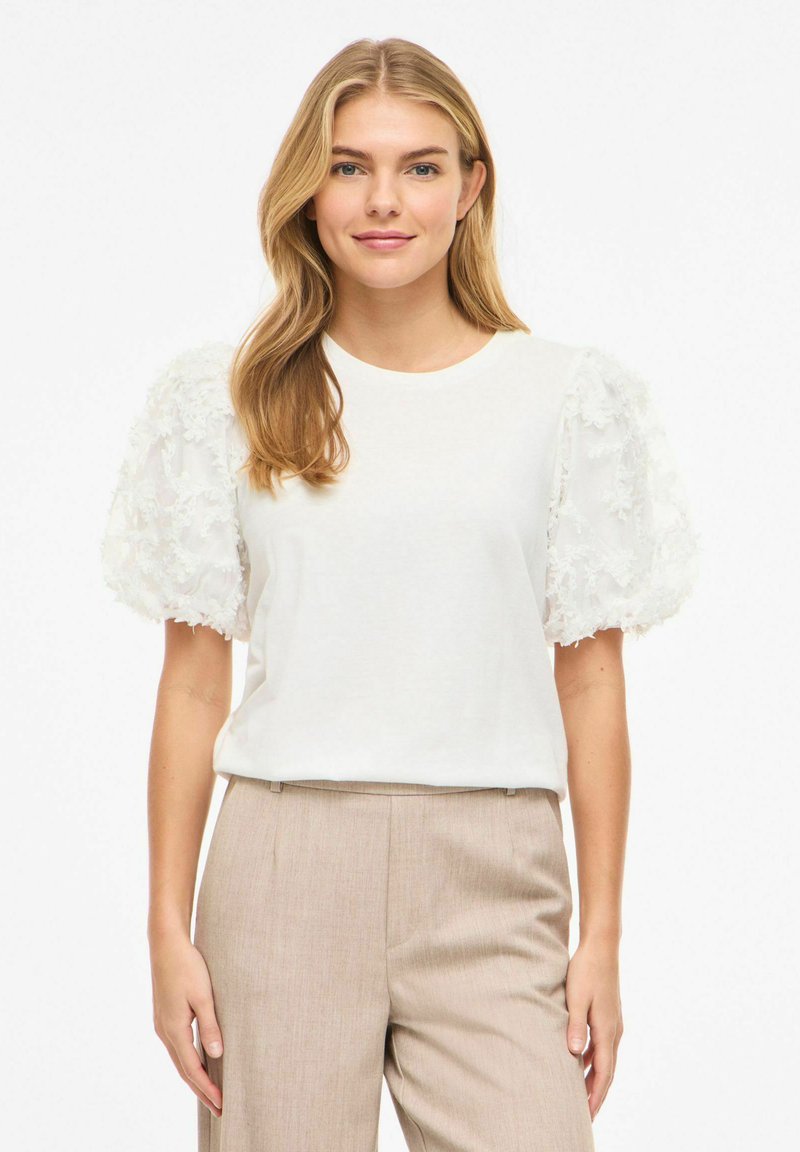 Femme blonde portant un haut blanc à manches bouffantes avec une texture florale et un pantalon beige taille haute, debout devant un fond blanc.