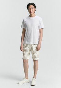 Weißes T-Shirt mit Rundhalsausschnitt kombiniert mit knielangen Shorts im palmenblätter Muster in Beige und Grün, getragen mit weißen Sneakers.